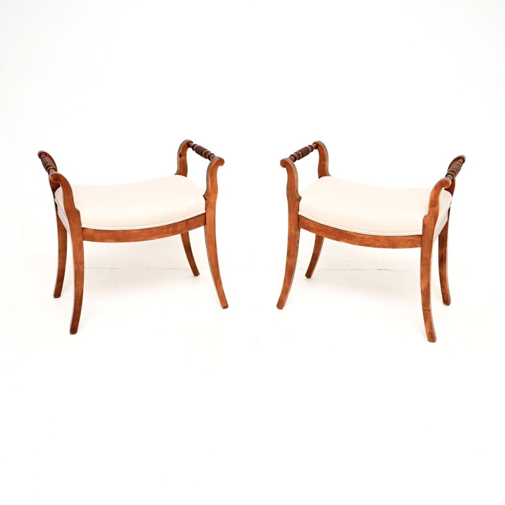 A pair of Swedish satin birch Biedermeier stools featuring curved cream upholstered seats, turned handle grips, and sweeping splayed legs.