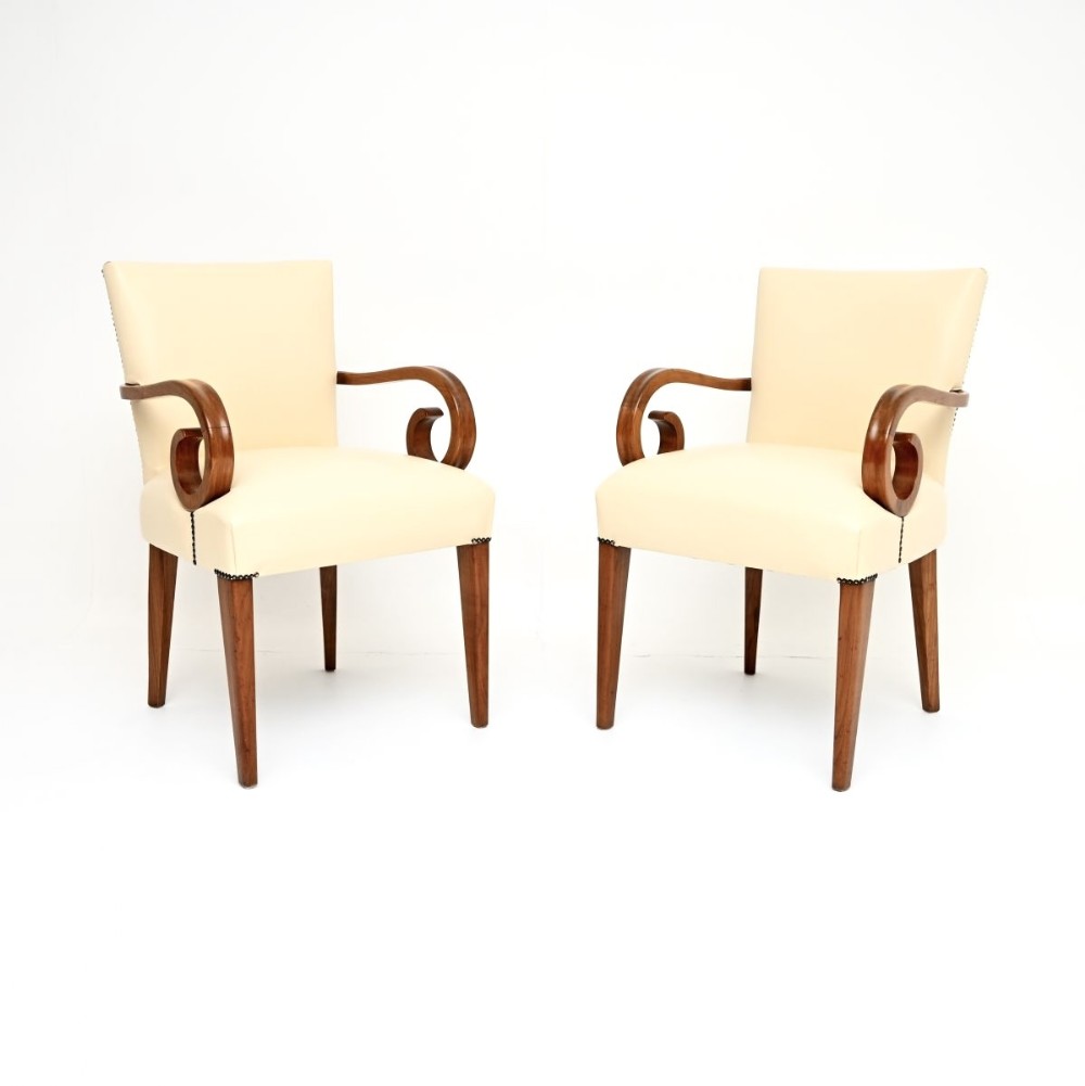 A pair of Art Deco armchairs featuring off-white leather upholstery with stud details and curved walnut arms on tapered legs.
