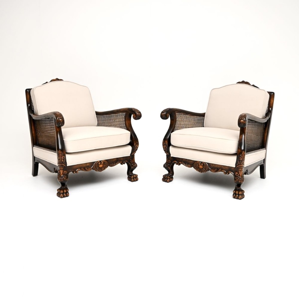 A pair of dark carved wood bergere armchairs featuring woven cane sides, paw feet, and light cream upholstered cushions.