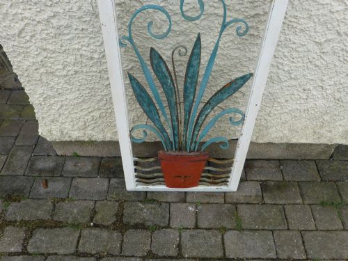 fig 1. A rectangular white metal frame housing a decorative wrought iron panel with a sunburst motif and stylised blue potted plant design.