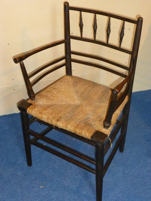 fig 1. An Arts and Crafts ebonised wooden armchair featuring turned back spindles, curved armrests, and a woven rush seat.