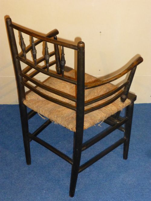 fig 2. An Arts and Crafts ebonised wooden armchair featuring turned back spindles, curved armrests, and a woven rush seat.