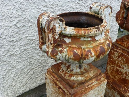 fig 2. A pair of Regency period cast iron campana urns with scrolled handles, displaying distressed off-white paint and surface rust, mounted on square cast iron pedestals.