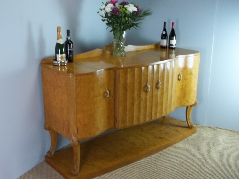 A burr walnut Art Deco sideboard featuring fluted central doors, curved side cupboards, brass handles, and cabriole legs resting on a solid plinth base.
