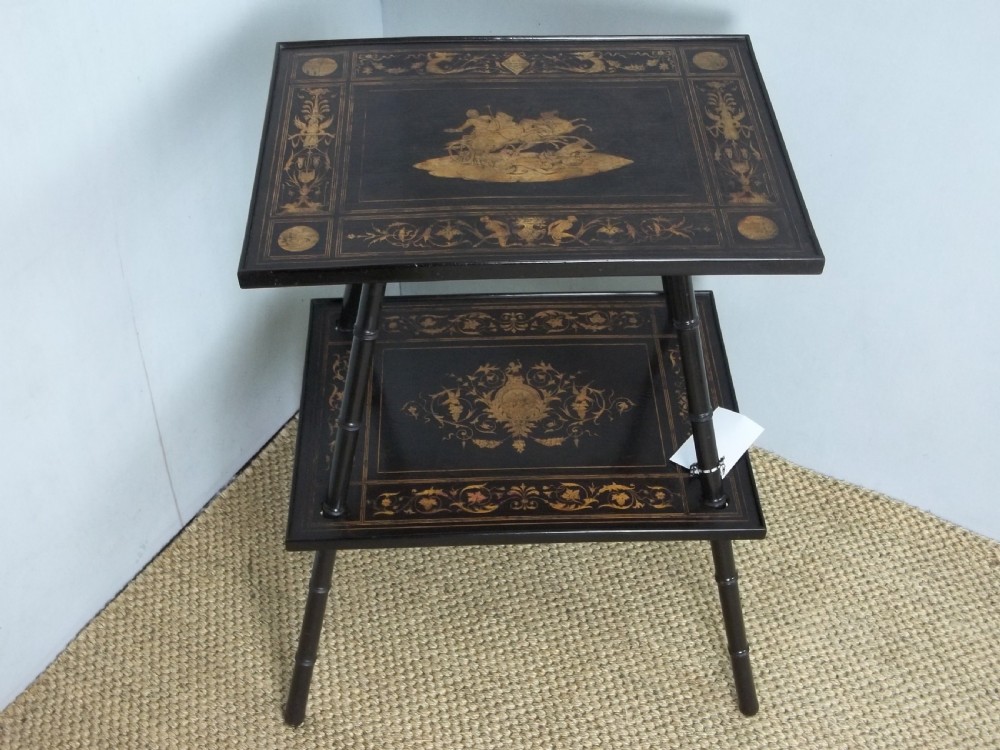 An ebonised two-tier side table displaying Sorrento marquetry inlay depicting a central chariot scene surrounded by decorative foliage and resting on turned legs.