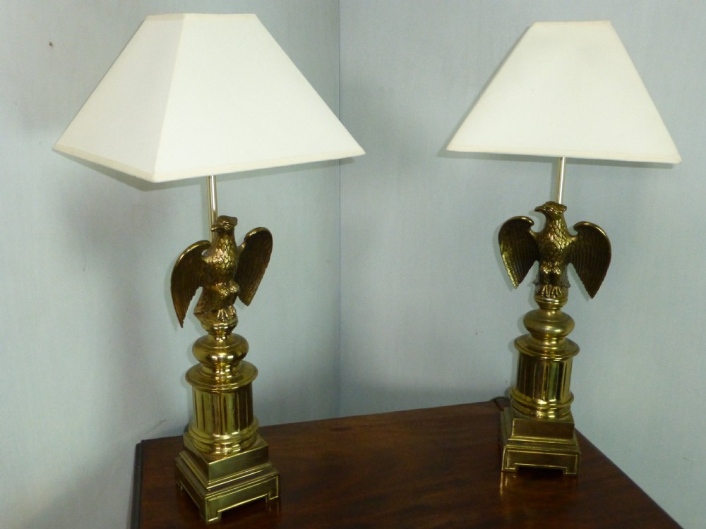 A pair of cast brass table lamps featuring eagle sculptures on stepped pedestal bases with cream pyramid shades.