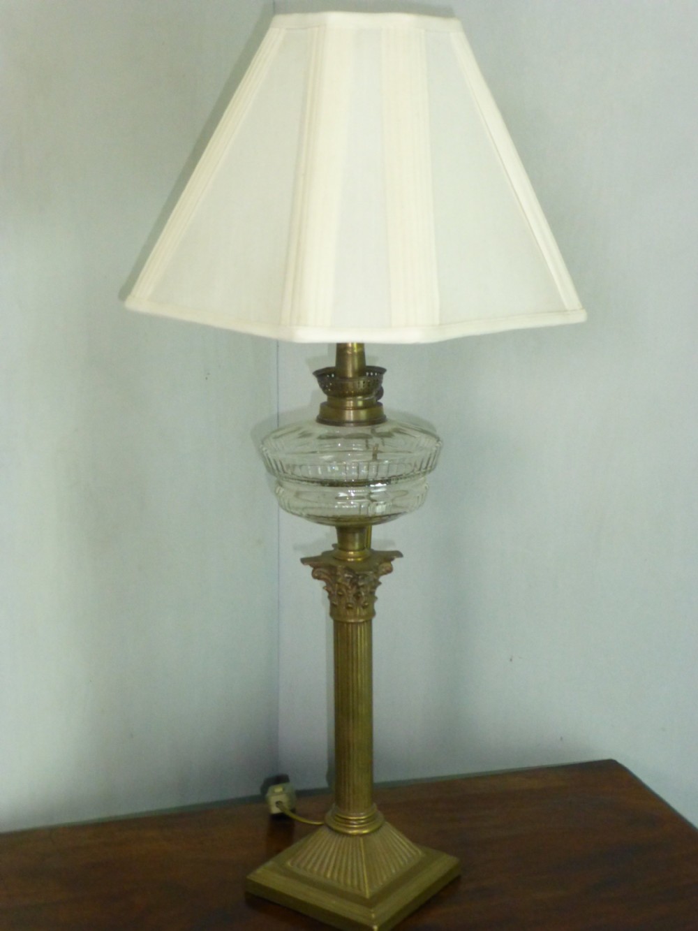 An antique Victorian brass table lamp featuring a fluted Corinthian column stem, a clear moulded glass reservoir, and a pleated hexagonal cream lampshade.
