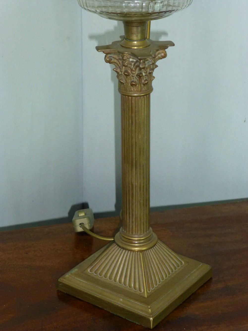 An antique Victorian brass table lamp featuring a fluted Corinthian column stem, a clear moulded glass reservoir, and a pleated hexagonal cream lampshade. View II