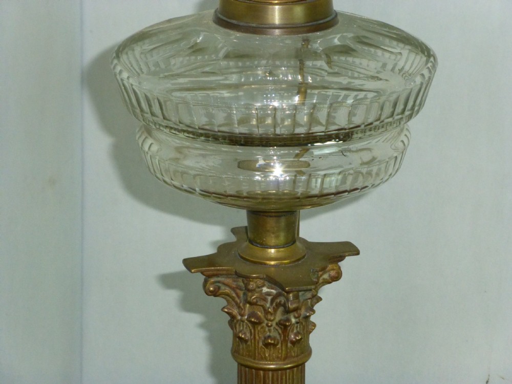 An antique Victorian brass table lamp featuring a fluted Corinthian column stem, a clear moulded glass reservoir, and a pleated hexagonal cream lampshade. View III