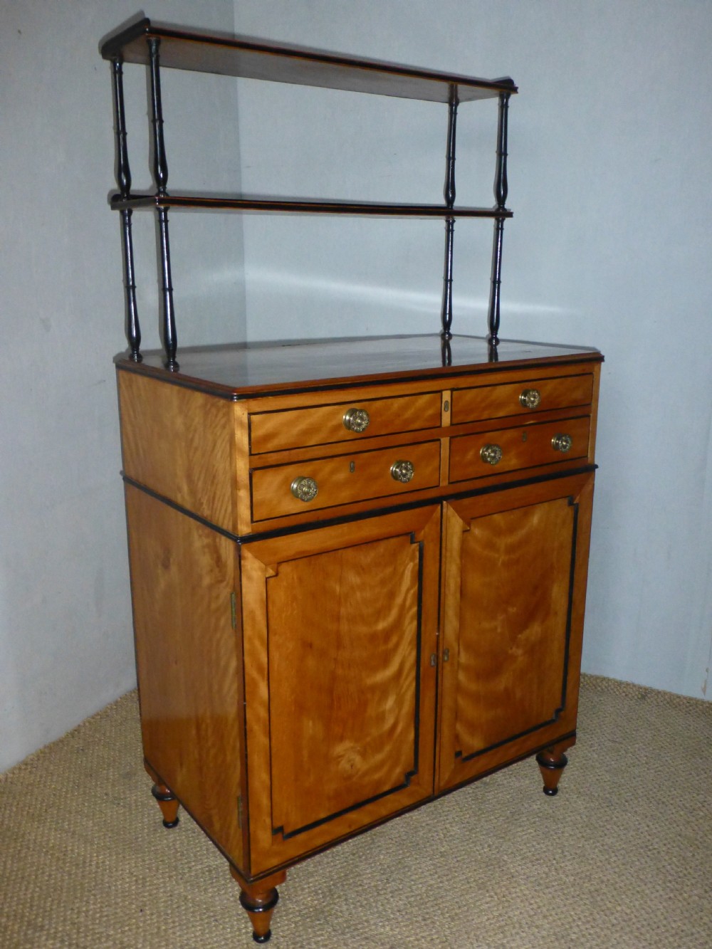 A Regency satinwood chiffonnier desk featuring two tiered upper shelves with ebonised turned supports above drawers and a double cupboard door cabinet.
