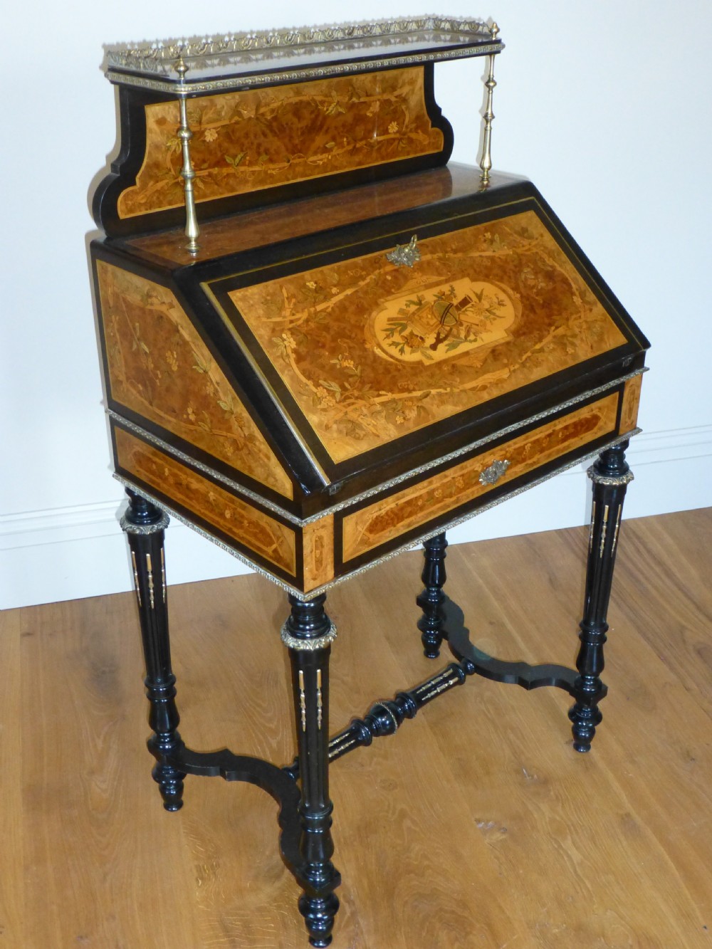 A nineteenth-century French slope-front writing desk featuring floral amboyna marquetry, ebonised turned legs with a shaped stretcher, and a brass gallery rail top.
