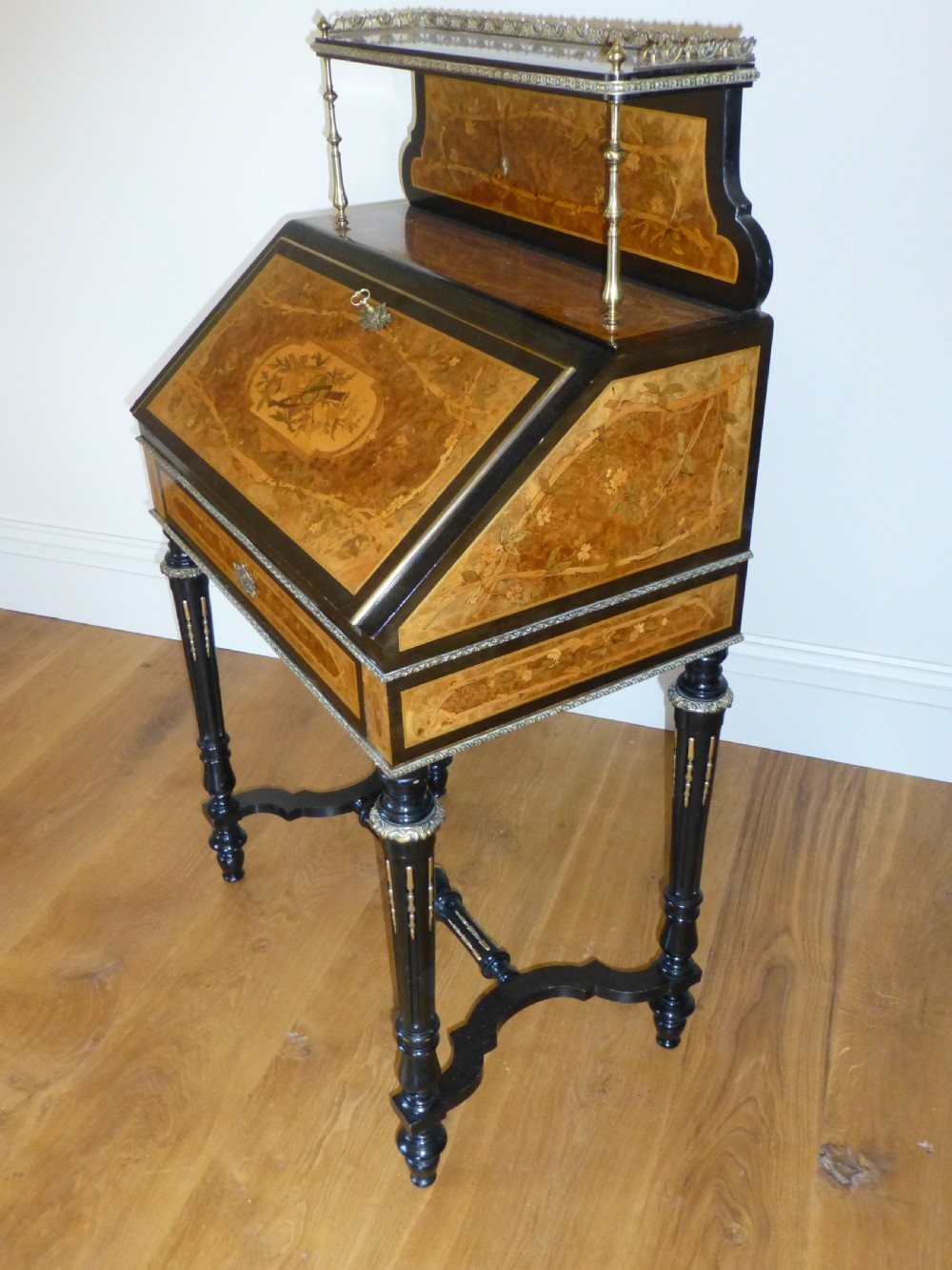 A nineteenth-century French slope-front writing desk featuring floral amboyna marquetry, ebonised turned legs with a shaped stretcher, and a brass gallery rail top. View XI