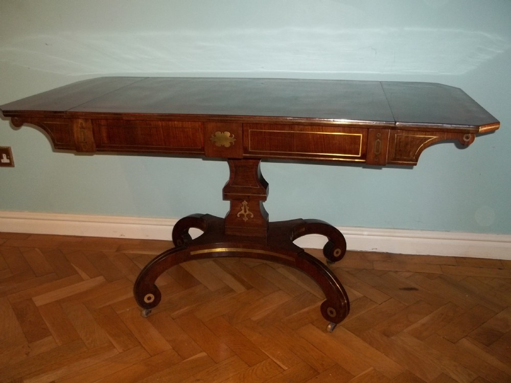 A Regency rosewood sofa table featuring drop leaves, brass inlay stringing, a central square pedestal, and splayed quadruped legs on castors.
