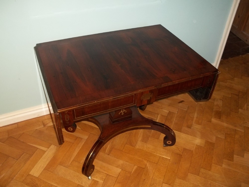 A Regency rosewood sofa table featuring drop leaves, brass inlay stringing, a central square pedestal, and splayed quadruped legs on castors. View II