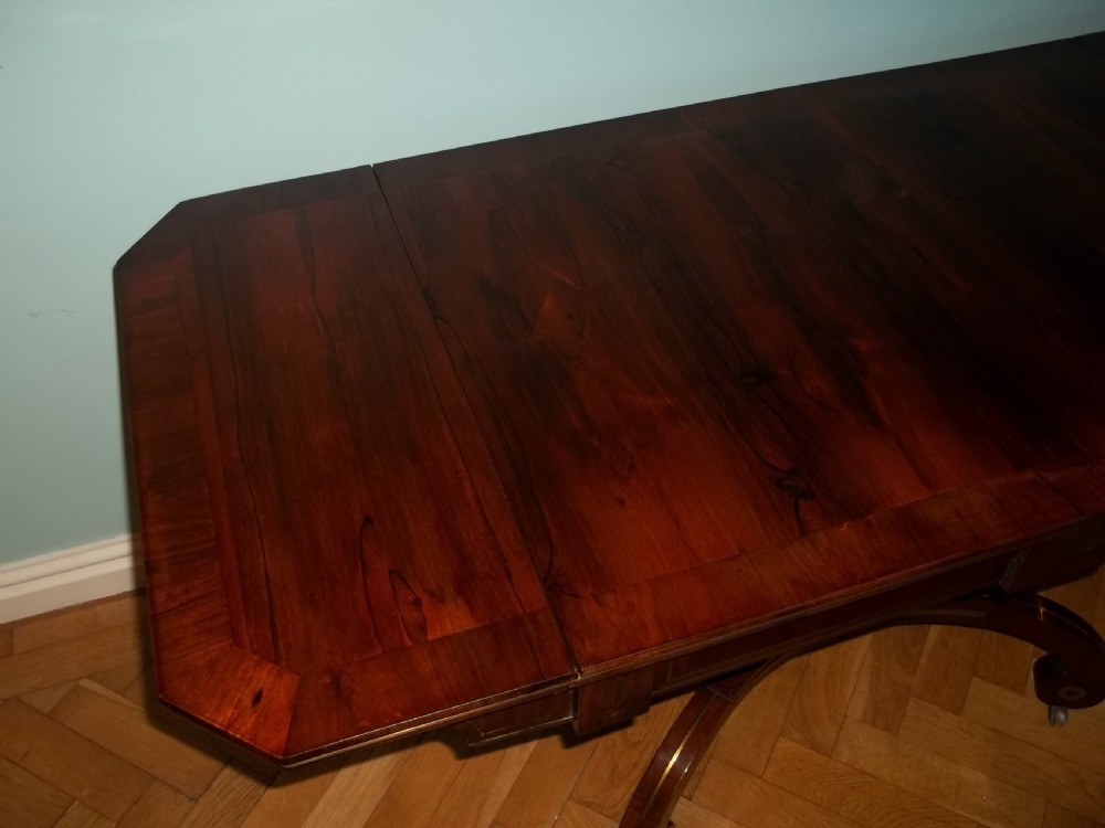 A Regency rosewood sofa table featuring drop leaves, brass inlay stringing, a central square pedestal, and splayed quadruped legs on castors. View III
