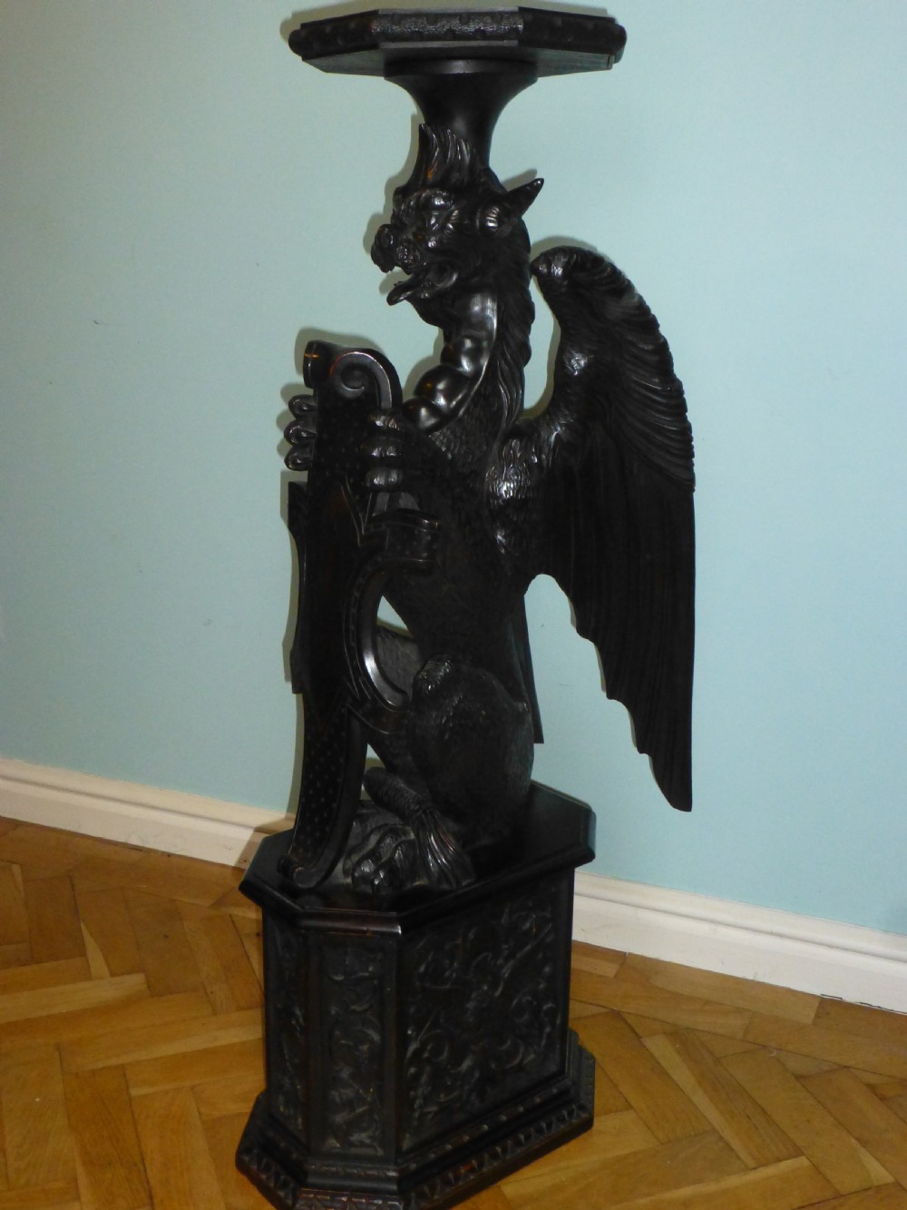 A dark carved wooden torchere stand featuring a detailed winged griffin holding a shield on an octagonal plinth base supporting an octagonal top.