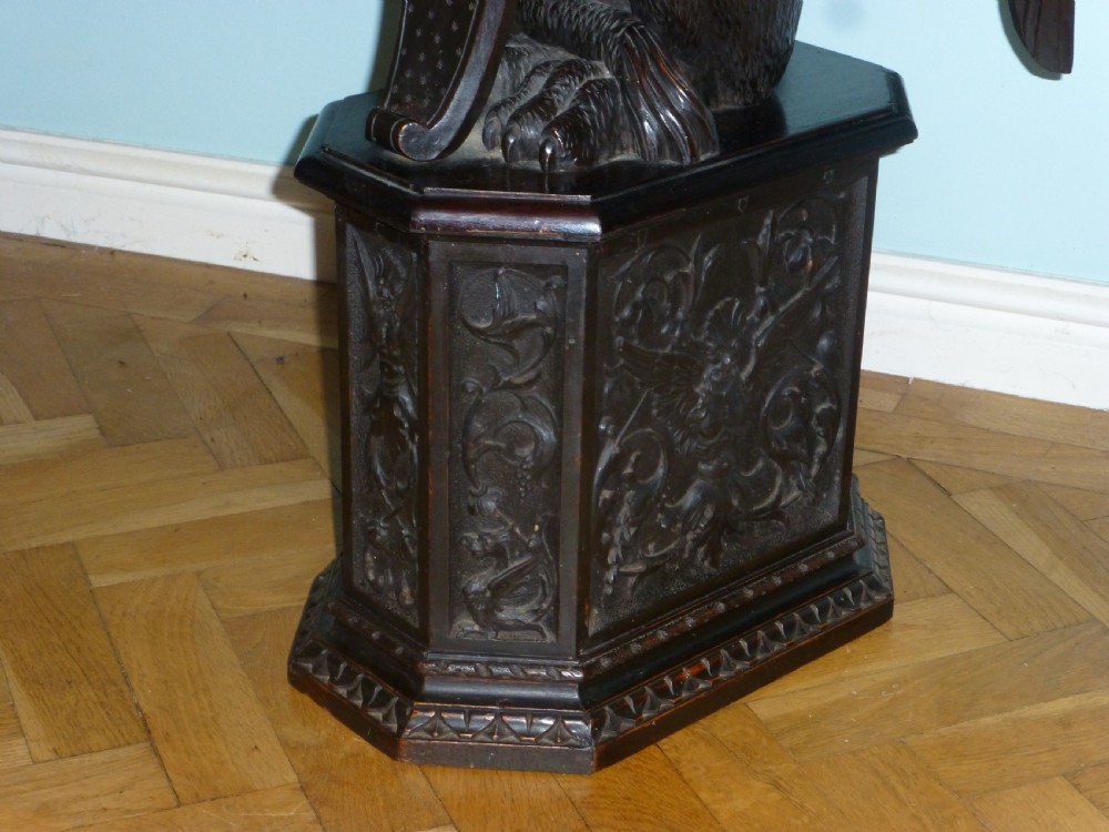 A dark carved wooden torchere stand featuring a detailed winged griffin holding a shield on an octagonal plinth base supporting an octagonal top. View II