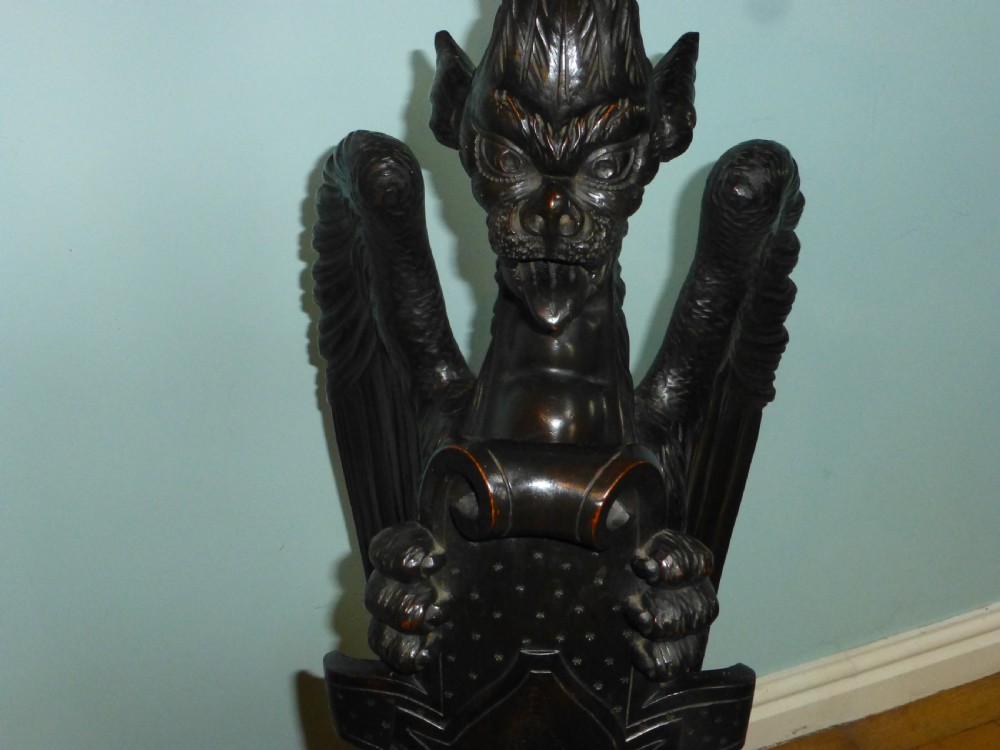 A dark carved wooden torchere stand featuring a detailed winged griffin holding a shield on an octagonal plinth base supporting an octagonal top. View III