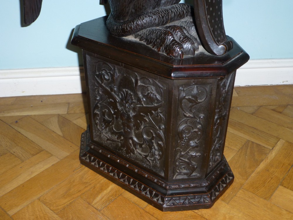 A dark carved wooden torchere stand featuring a detailed winged griffin holding a shield on an octagonal plinth base supporting an octagonal top. View IV