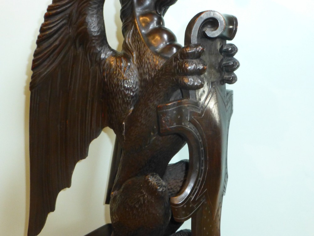 A dark carved wooden torchere stand featuring a detailed winged griffin holding a shield on an octagonal plinth base supporting an octagonal top. View V
