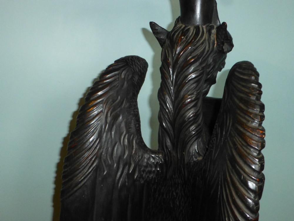 A dark carved wooden torchere stand featuring a detailed winged griffin holding a shield on an octagonal plinth base supporting an octagonal top. View VI