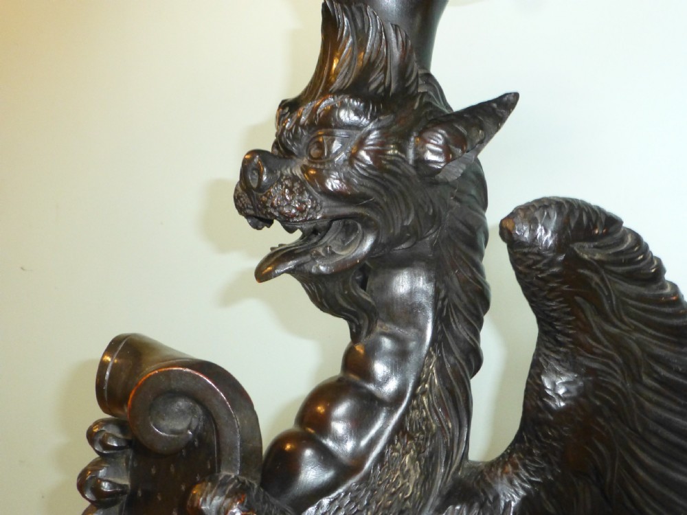 A dark carved wooden torchere stand featuring a detailed winged griffin holding a shield on an octagonal plinth base supporting an octagonal top. View X