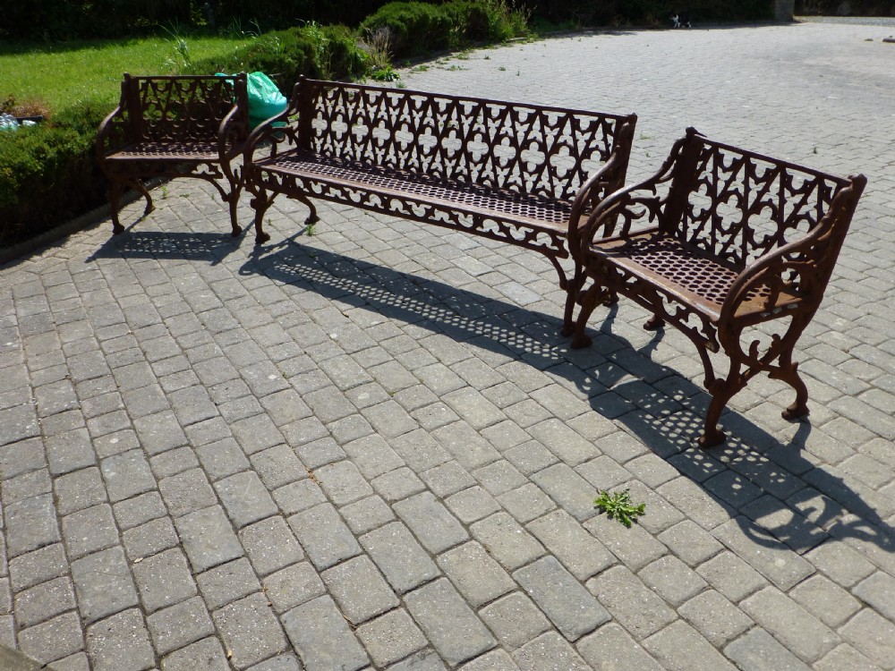 A three-piece antique cast iron garden set comprising a wide bench and two matching armchairs featuring openwork Gothic tracery backrests and weathered brown surface oxidisation.