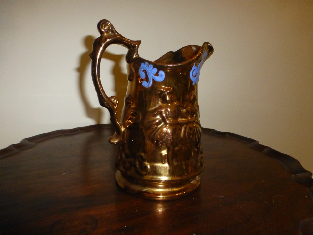 A Victorian Staffordshire earthenware jug featuring a reflective copper lustre glaze, moulded relief figures, blue floral scroll decoration, and a shaped handle. View II