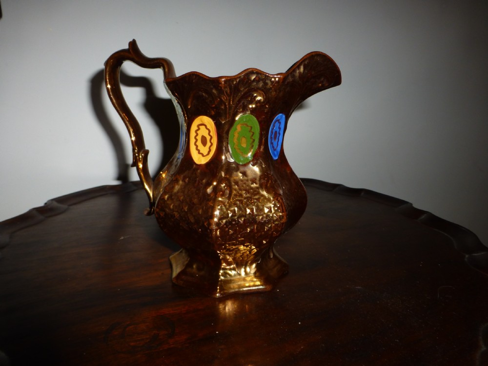 A Victorian copper lustre ceramic milk jug featuring a scalloped rim, scroll handle, and hand-painted enamel oval motifs in yellow, green, and blue.