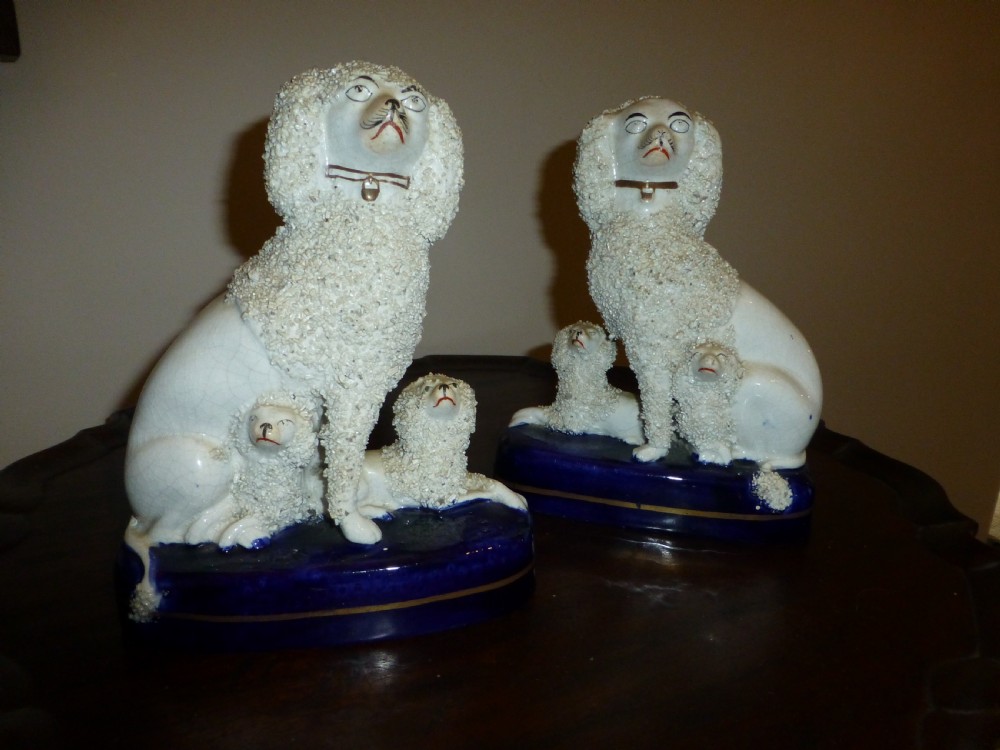 A pair of white textured Victorian Staffordshire ceramic poodles seated on cobalt blue bases with two small puppies beside each.