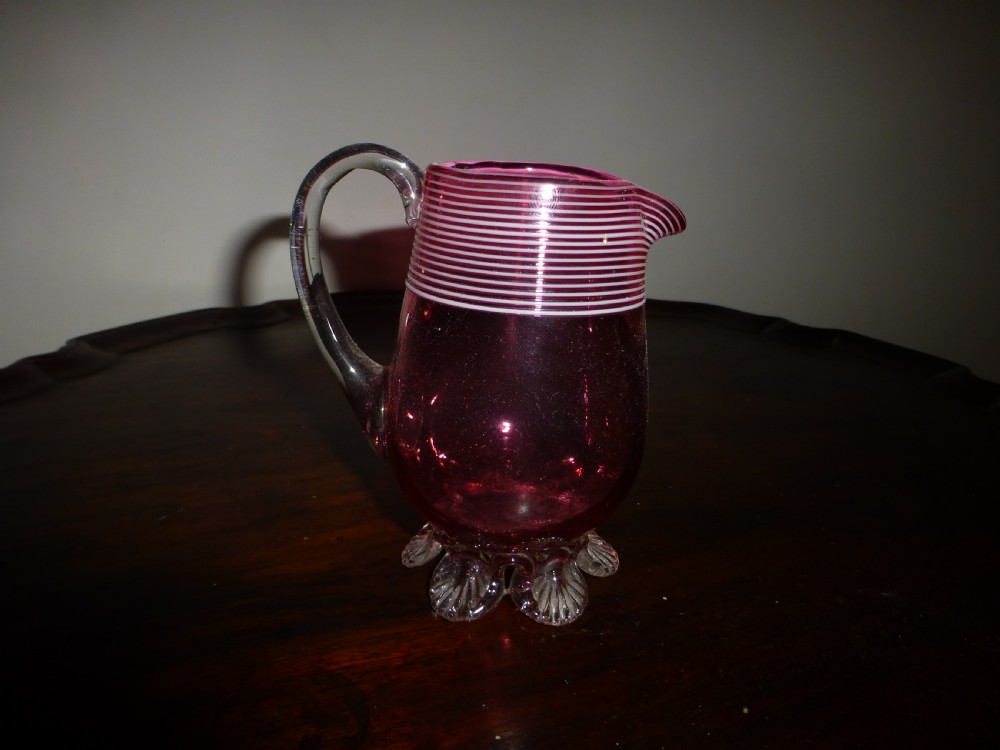 A Victorian cranberry glass cream jug featuring white threaded piping around the rim, a clear applied handle, and clear shell feet.
