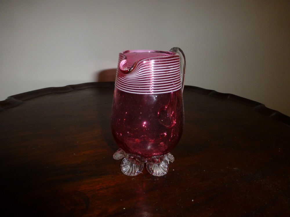 A Victorian cranberry glass cream jug featuring white threaded piping around the rim, a clear applied handle, and clear shell feet. View II