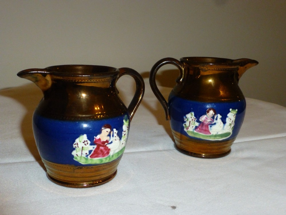 A pair of small ceramic cream jugs featuring metallic copper lustre glaze on the rims and bases with cobalt blue central bands showing hand-painted figures.