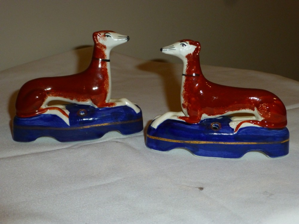 A pair of Victorian Staffordshire ceramic inkwells formed as reclining rust-red greyhounds set upon cobalt-blue glaze bases with gilded trim.
