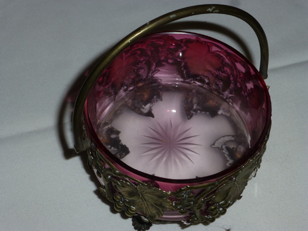A Victorian cranberry glass preserve dish set inside an ornate metal basket with a swing handle and vine leaf detailing. View II