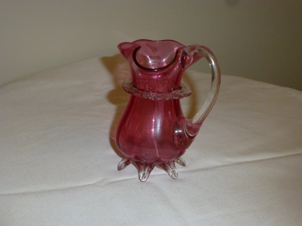 A Mid-Victorian cranberry glass jug featuring a crimped rim, an applied clear glass handle, a decorative clear collar, and clear feet. View II