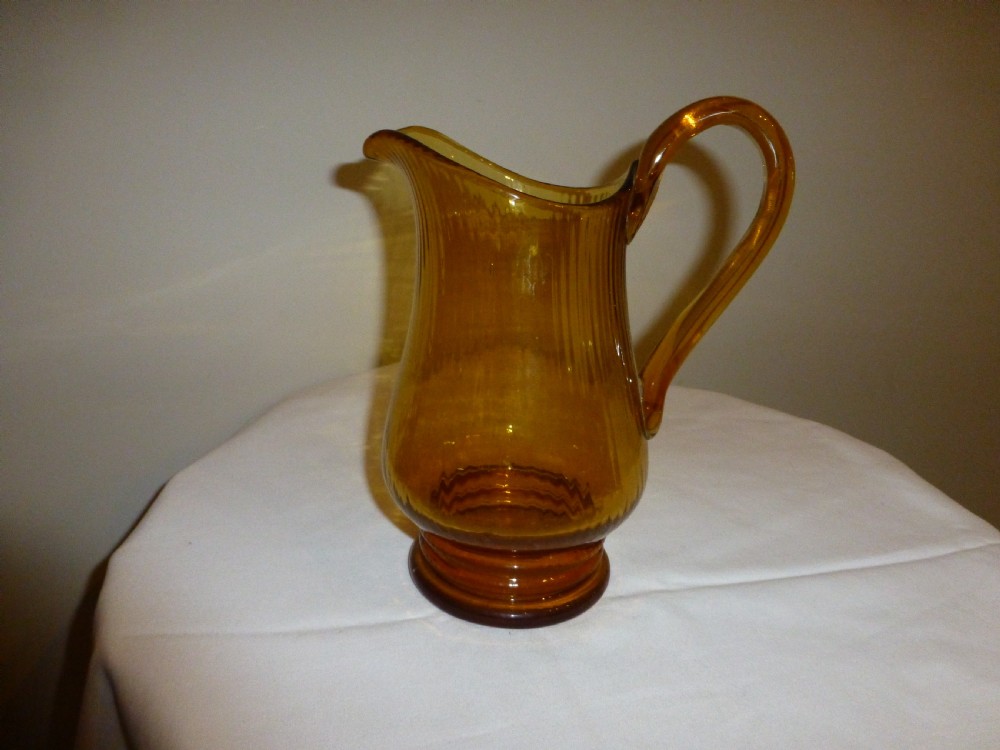 An amber glass lemonade jug featuring a ribbed body, an applied looped handle, a flared spout, and a solid circular base.