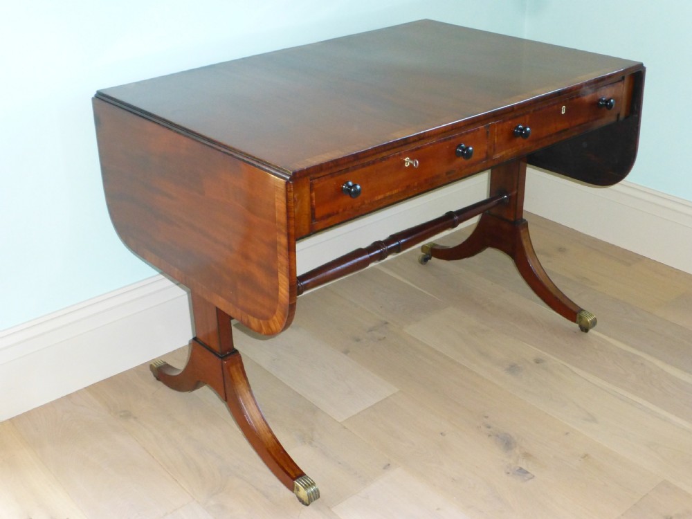 A Regency mahogany drop-leaf sofa table featuring two drawers with dark round knobs, splayed legs ending in brass castors, and a turned central stretcher.