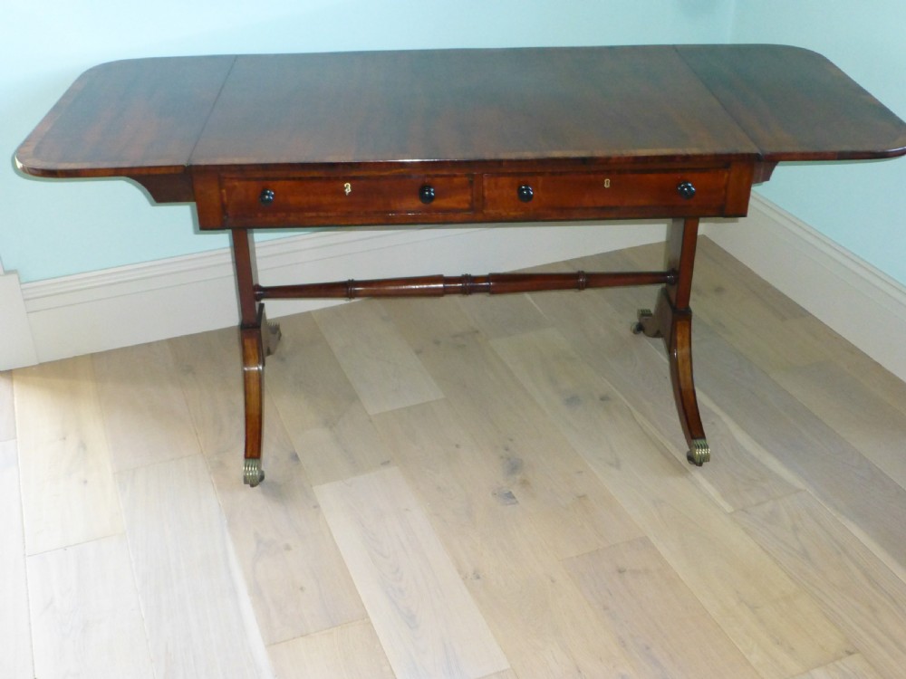 A Regency mahogany drop-leaf sofa table featuring two drawers with dark round knobs, splayed legs ending in brass castors, and a turned central stretcher. View V