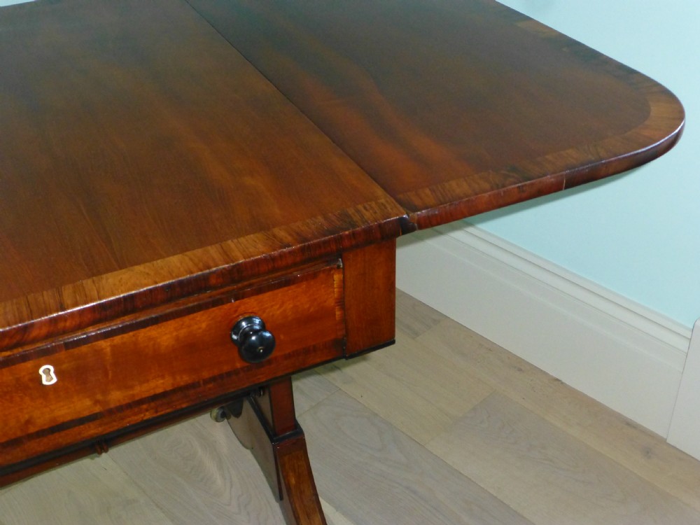 A Regency mahogany drop-leaf sofa table featuring two drawers with dark round knobs, splayed legs ending in brass castors, and a turned central stretcher. View VII