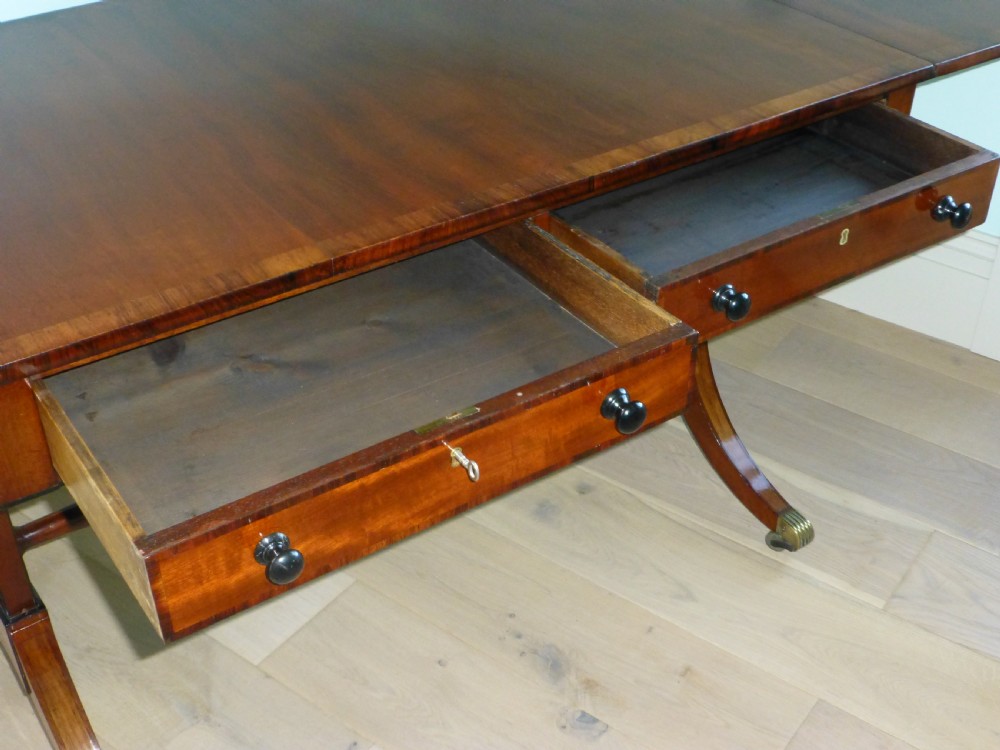 A Regency mahogany drop-leaf sofa table featuring two drawers with dark round knobs, splayed legs ending in brass castors, and a turned central stretcher. View VIII