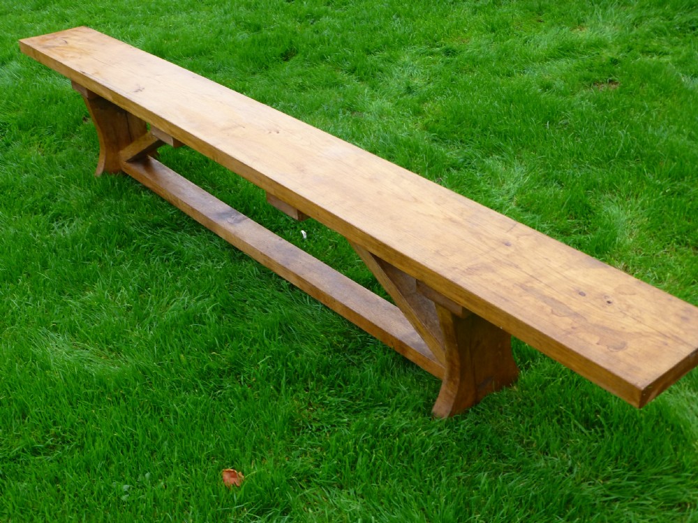 A long, solid oak trestle bench featuring a single thick wooden seat supported by shaped end trestles and a low central stretcher rail.