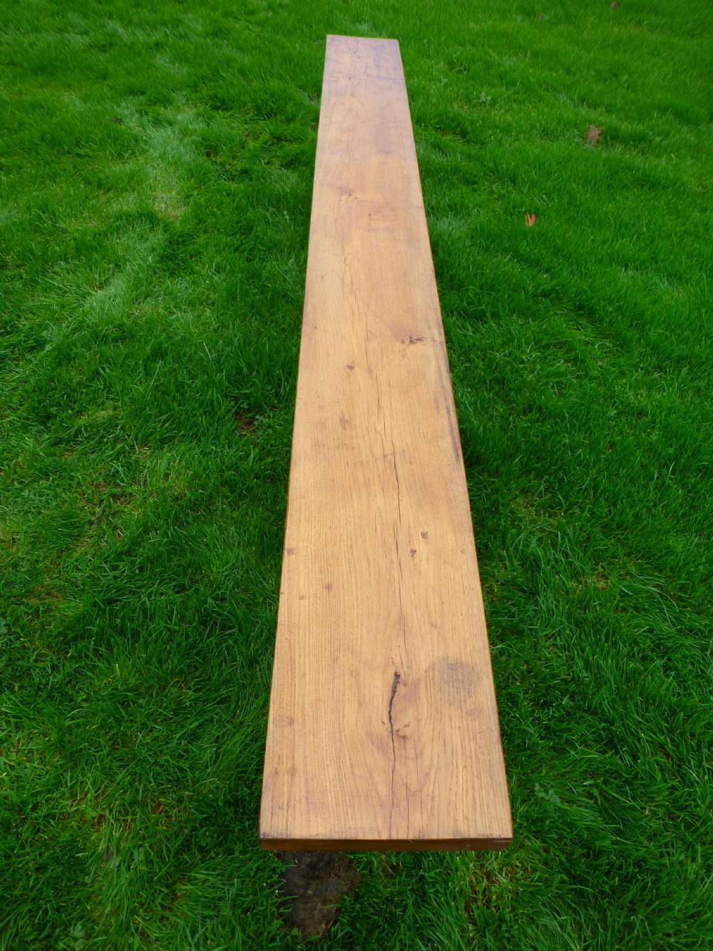 A long, solid oak trestle bench featuring a single thick wooden seat supported by shaped end trestles and a low central stretcher rail. View II