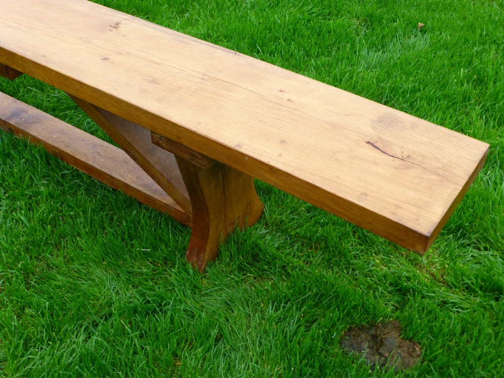 A long, solid oak trestle bench featuring a single thick wooden seat supported by shaped end trestles and a low central stretcher rail. View III