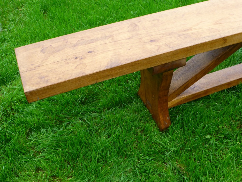 A long, solid oak trestle bench featuring a single thick wooden seat supported by shaped end trestles and a low central stretcher rail. View V