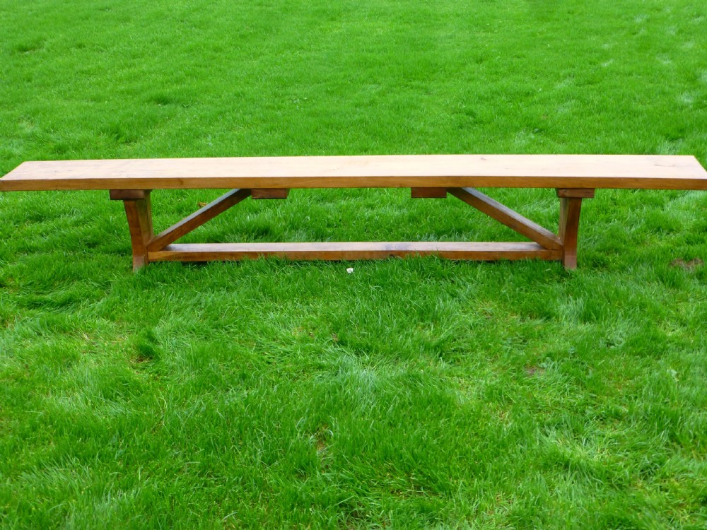A long, solid oak trestle bench featuring a single thick wooden seat supported by shaped end trestles and a low central stretcher rail. View VI