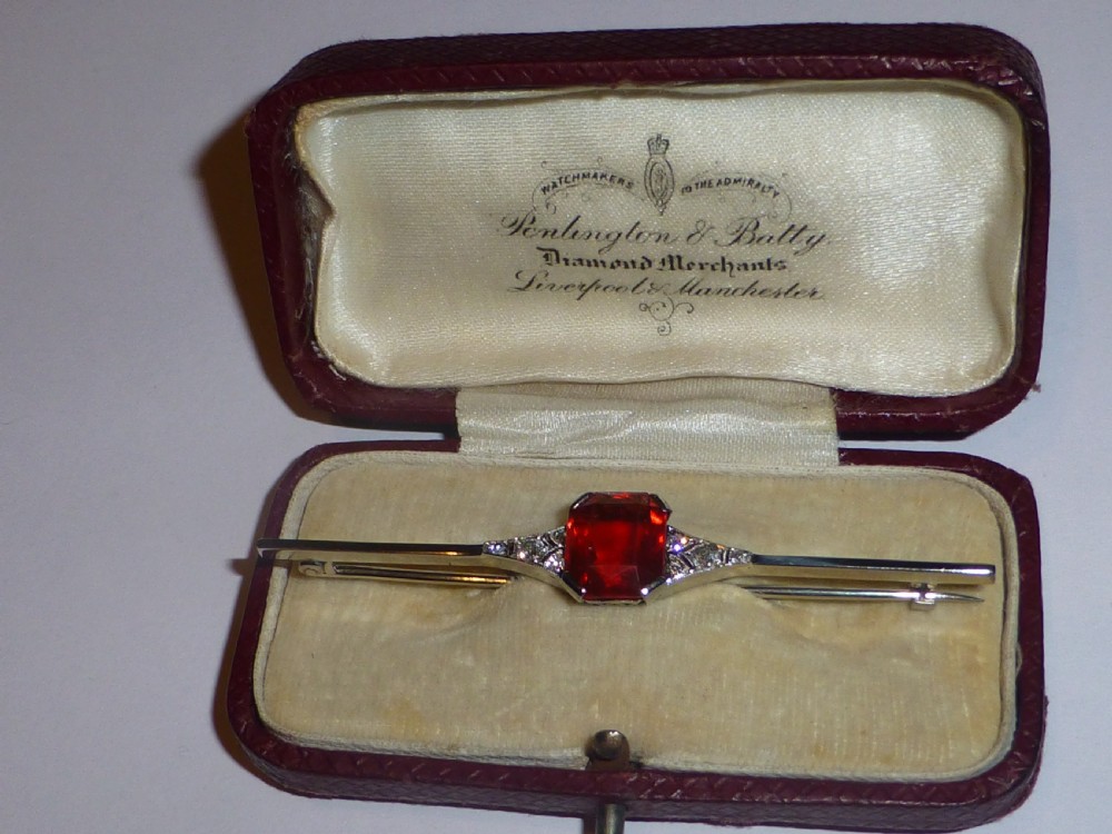 An Edwardian 18ct white gold bar brooch featuring a central emerald-cut orange fire opal flanked by diamond accents. View II