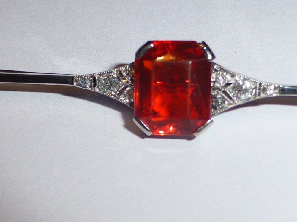 An Edwardian 18ct white gold bar brooch featuring a central emerald-cut orange fire opal flanked by diamond accents. View V