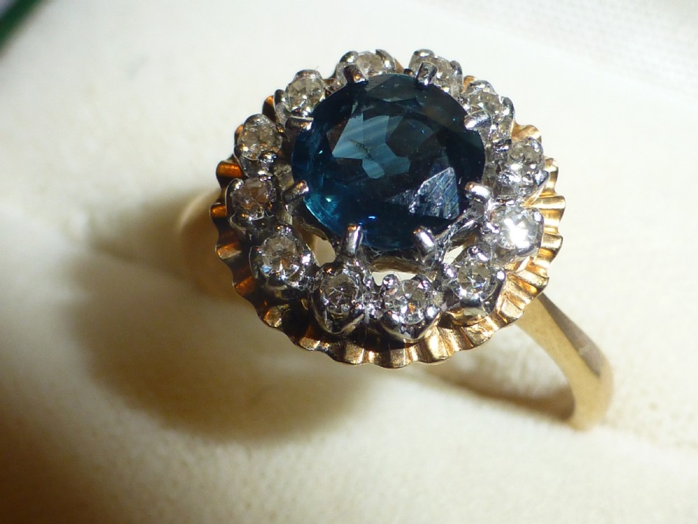 An antique gold ring featuring a central round deep blue sapphire framed by a halo of small diamonds within a scalloped edge setting. View II
