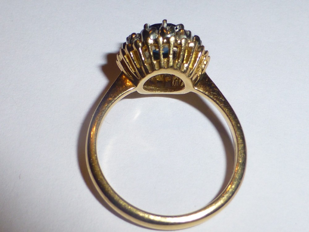 An antique gold ring featuring a central round deep blue sapphire framed by a halo of small diamonds within a scalloped edge setting. View V